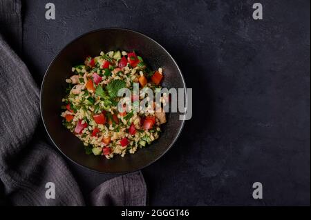 Tradizionale insalata libanese tovouleh fatto di bulgur o couscous, carne di pollame, prezzemolo, menta in ciotola scura con tovagliolo su sfondo nero. Vista dall'alto, spazio di copia Foto Stock