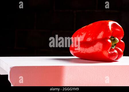 Fotografia di un peperone rosso bagnato e adagiato su una panca bianca e uno sfondo nero. La foto è scattata in formato orizzontale e ha spazio da mettere Foto Stock