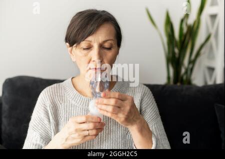 Vista laterale una donna anziana sta usando l'inalatore per il trattamento di flu e freddo seduto sul divano a casa. Concetto di sanità e medicina Foto Stock