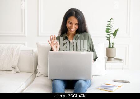 Videoconferenza, videoconnessione con i colleghi da casa. Giovane donna indiana che indossa una maglietta casual utilizzando un computer portatile per comunicare online, ondeggiandosi in webcam, salutando i partecipanti al webinar Foto Stock