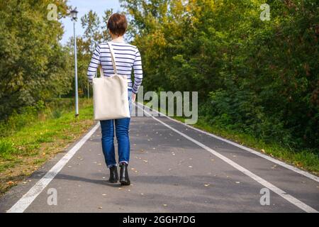 Donna che porta una borsa per la spesa vuota riutilizzabile che cammina nel mockup del parco. Borsa tote eco-compatibile con tele da tenuta a mano femmina Foto Stock