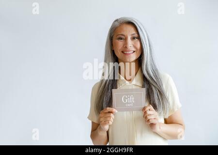 Sorridente donna asiatica dai capelli grigi tiene la carta regalo in piedi su sfondo chiaro Foto Stock