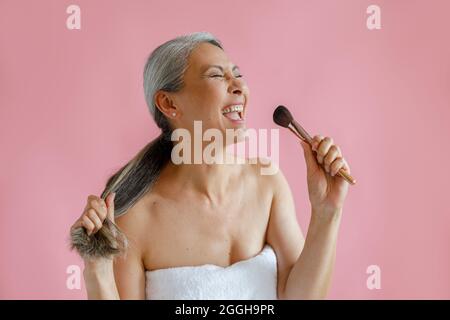 Jouful donna asiatica di mezza età canta usando spazzola cosmetica come microfono su sfondo rosa Foto Stock