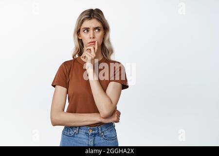 Immagine di una donna preoccupata che guarda da parte esitante, pensando a qualcosa, in piedi su sfondo bianco Foto Stock