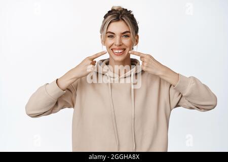 Clinica dentale, concetto di stomatologia. Sorridendo bella ragazza punta le dita ai suoi denti sani bianchi, sorriso perfetto, si leva sullo sfondo dello studio Foto Stock