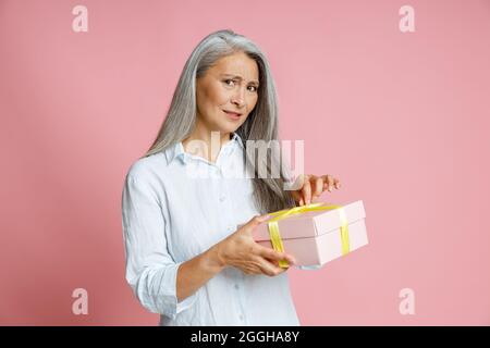 Dubiting grigio capelli Asian donna apre scatola regalo posa su sfondo rosa in studio Foto Stock