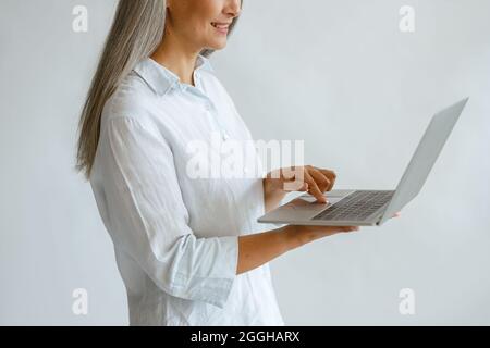 Donna con capelli hoary in blusa bianca usa il computer portatile su sfondo chiaro in studio Foto Stock