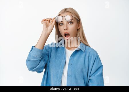 Immagine della ragazza bionda sorpresa decolga i suoi occhiali, la mascella goccia e lo stare impressionato alla macchina fotografica, in piedi su sfondo bianco Foto Stock