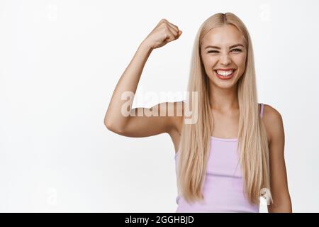 Allegra donna bionda che canta, canta fisti e sorridente felice, raduno per la squadra, festeggiamenti, in piedi su sfondo bianco Foto Stock