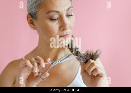 La donna asiatica dubitante taglia le estremità sfilacciate dei capelli lunghi grigi su sfondo rosa in studio Foto Stock