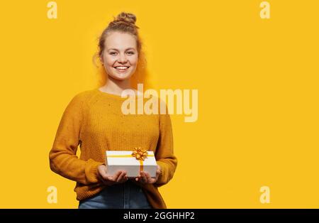 Sorridendo bella ragazza bionda che tiene una scatola del regalo isolata sopra lo sfondo giallo. Buon compleanno, Natale, San Valentino concetto. Luogo per Foto Stock