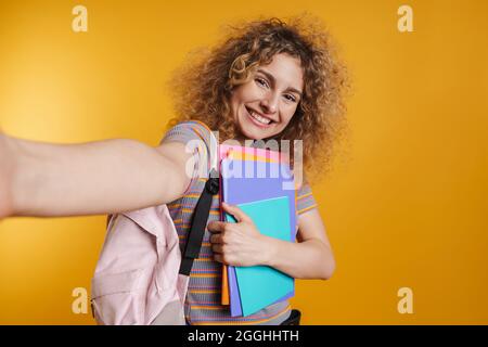 Giovane studentessa felice con zaino in piedi che tiene libri di testo su sfondo giallo parete, prendendo un selfie Foto Stock