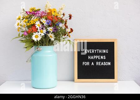 Tutto accade per un motivo. Citazione motivazionale sulla lettera e bouquet fiori colorati su tavola bianca contro parete di pietra grigia. Concetto ispi Foto Stock