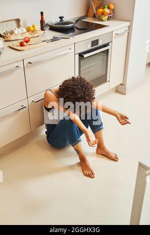 Crestcaduto solo ricci-capelli giovane afroamericana donna seduta a piedi nudi sul pavimento in cucina Foto Stock