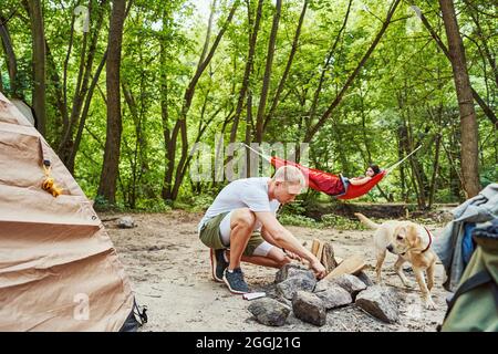 Il giovane uomo sta facendo il fuoco vicino alla tenda mentre la sua ragazza è sdraiata e rilassante in amaca nella foresta Foto Stock