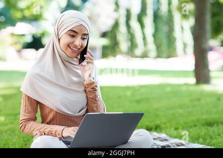 Felice attraente giovane medio orientale femmina in hijab guarda in notebook, chiamata per telefono, lavora a distanza nel parco Foto Stock