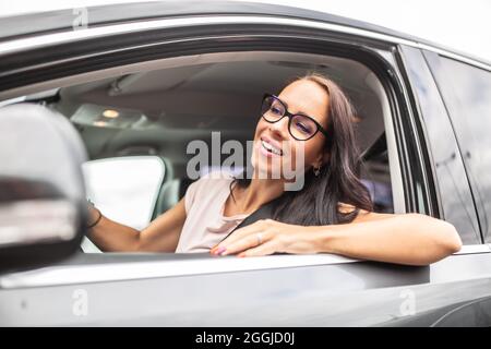 Felice driver femmina in occhiali guarda attraverso il finestrino laterale aperto di una macchina che guida. Foto Stock