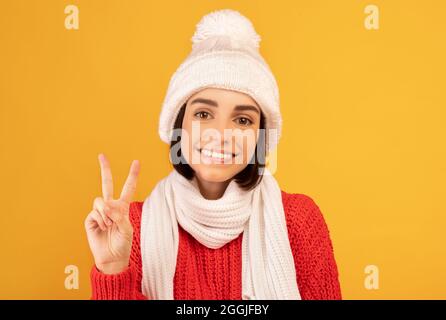 Donna felice in caldo maglione e cappello lavorato a maglia sorridente e facendo gesto di pace su sfondo giallo studio Foto Stock