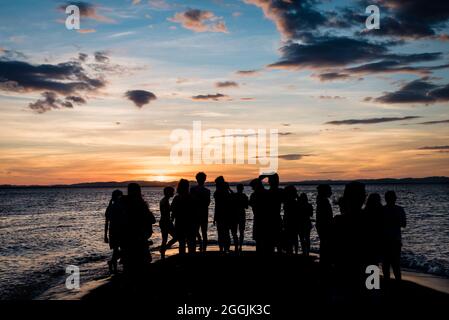 Silhouette di un gruppo di giovani con le spalle girate guardando il tramonto sulla riva con un cielo colorato con le nuvole Foto Stock