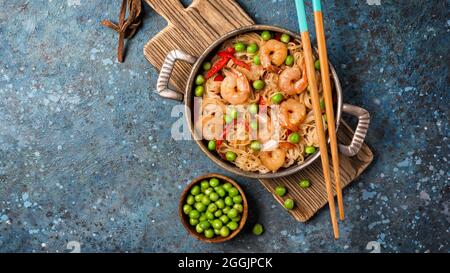 Vista dall'alto della padella con spaghetti di riso cinesi, gustosi gamberi, piselli verdi in ciotola e pepe rosso su tavola di legno e sfondo blu in cemento Foto Stock