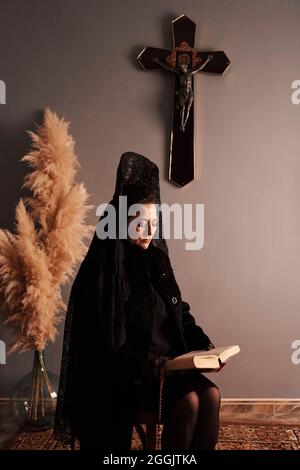 Una donna vestita di nero si siede a leggere una bibbia di fronte ad un crocifisso Foto Stock