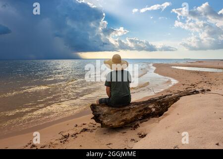 Uomo in cappello siediti sul tronco dell'albero guardando la tempesta e il tramonto sulla spiaggia del fiume Tapajos nella foresta pluviale di Amazzonia. Concetto di natura, conservazione, ambiente. Foto Stock
