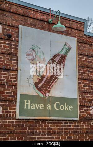 Un vecchio cartello pubblicitario di Coca-Cola d'epoca attaccato ad una parete esterna all'esterno sotto una luce su un edificio in mattoni indossato e sbiadito di un holdi giovane Foto Stock