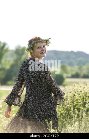 Giovane donna con una corona di fiori naturali sulla testa e fiori in mano corre vivace tra campi Foto Stock