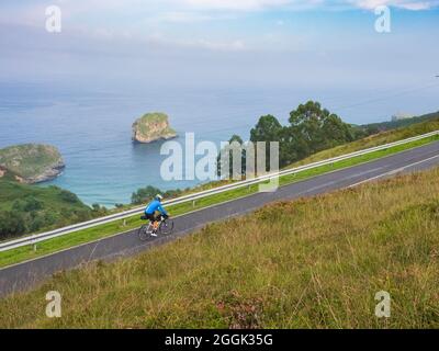 Tour in bici con bagagli, bikepacking su una strada costiera solitaria nelle Asturie, Spagna settentrionale con vista sull'Oceano Atlantico e la spiaggia di Playa de la Ballota vicino a Llanes nelle Asturie Foto Stock