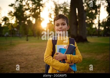 Ritratto sicuro di un felice bel ragazzo della scuola 9 anni con borsa della scuola che tiene i libri di lavoro e la cassa della matita nelle mani e cute che sorride posando Foto Stock