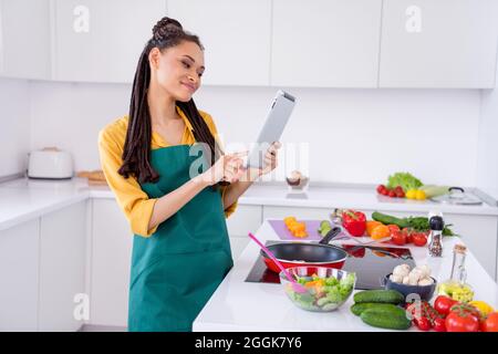 Foto di adorabile dolce pelle scura donna vestita camicia gialla cucina lettura dispositivo moderno interno casa casa camera Foto Stock