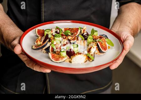 Persone al lavoro. Le mani dello chef stanno tenendo un piatto con insalata estiva con fichi maturi, formaggio di capra chevre, rotolo di cetrioli freschi, barbabietola e senape l Foto Stock