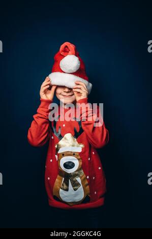 Studio girato di un ragazzo che indossa un cappello e un pullover di natale facendo il divertimento di fronte a uno sfondo blu Foto Stock
