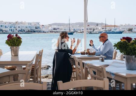 Città di Mykonos, Grecia - 24 settembre 2019: Coppia seduta e parlare ai tavoli all'aperto di un ristorante vicino all'acqua in Hora (noto anche come Mykono Foto Stock