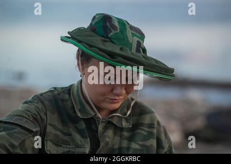 Una giovane donna in abiti camouflage costruisce un fuoco sulla riva del fiume. Tempo di tramonto. Foto Stock