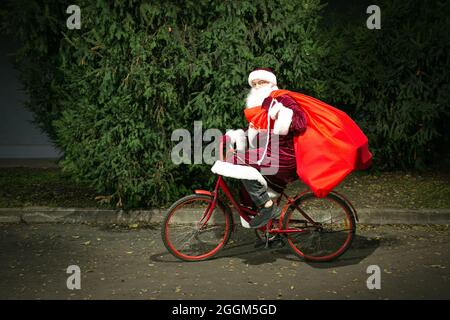 Babbo Natale corre una bicicletta e porta una grande borsa di regali. Primo piano sullo sfondo di alberi di conifere. Foto Stock