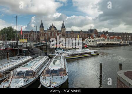 Amsterdam, Paesi Bassi - 14 agosto 2021: Gruppo di barche turistiche sul canale ormeggiate di fronte alla stazione ferroviaria rossa e dorata di Centraal, sotto il paesaggio blu nuvoloso. Foto Stock