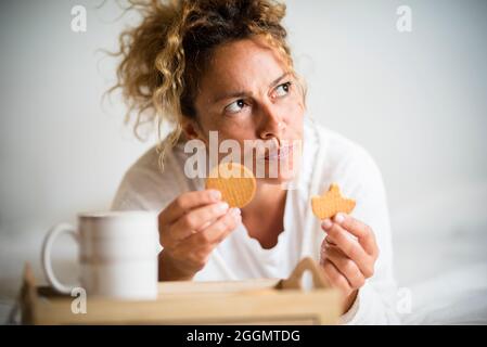 Donna adulta preoccupata con biscotti e caffè al mattino colazione a casa stesa a letto - donna di mezza età piuttosto con cibo e nutrizione pensieri uno Foto Stock