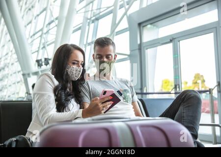Coppia in viaggio in maschera controllare ultimo aggiornamento su un telefono cellulare durante l'attesa in un terminal aeroporto. Foto Stock
