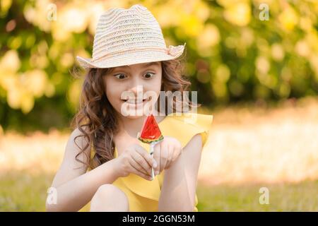 ragazza in abiti gialli e un cappello si siede sull'erba nel parco in estate con un lecca in forma di una fetta di cocomero. Foto di alta qualità Foto Stock