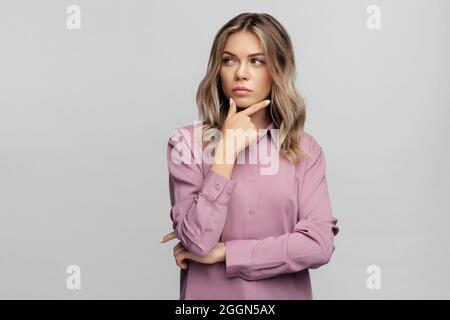 Pensiero giovane donna pensante premuroso guardare da parte braccio di sostegno sul mento confuso isolato sopra il muro grigio Foto Stock