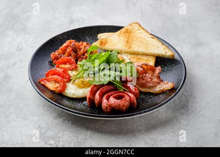 Uova fritte con salsiccia, pancetta, pomodori e fagioli su un piatto con toast Foto Stock
