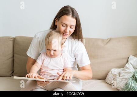 Ragazza curiosa del bambino seduta sulle ginocchia della madre e libro di lettura con le immagini. Mamma sorridente. Spazio di copia Foto Stock