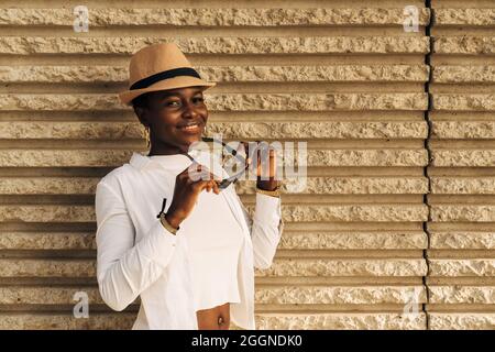 Sorridente donna afroamericana che indossa un cappello di paglia e tiene gli occhiali da sole mentre si posa all'aperto. Foto Stock