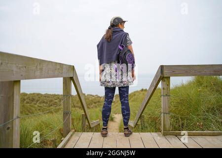 Vista posteriore di una giovane donna che guarda verso la spiaggia e il mare di fronte a lei in una giornata trascorsa in Galles Foto Stock