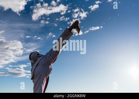 Ragazzo irriconoscibile che pratica arti marziali in bianco kimono all'aperto con uno sfondo di cielo con le nuvole in una giornata di sole. Concetto di karate. Foto Stock