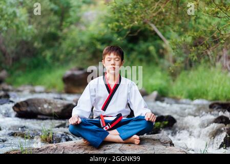 Ritratto di un ragazzo giapponese taekwondo seduto in natura e fare esercizi di meditazione. Il giovane asiatico nero cintura karate sitter. Foto Stock