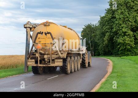 Un trattore con un moderno rimorchio per fertilizzanti che corre su strada di campagna lungo il campo in una giornata estiva. Foto Stock