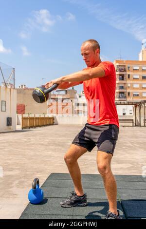 Giovane caucasico che fa squat con pesi campana sulla strada, si allena per mettersi in forma e sentirsi bene sul suo corpo. Concetto di vita sana Foto Stock