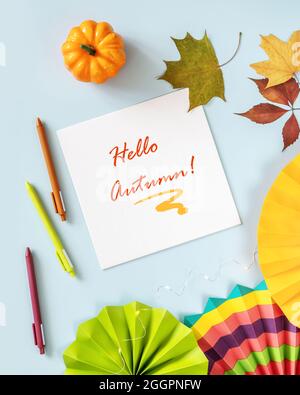 Luce, composizione autunnale con foglio di carta bianco con iscrizione Hello Autunno, ventilatori colorati, penne e decorazioni su sfondo blu. A c Foto Stock